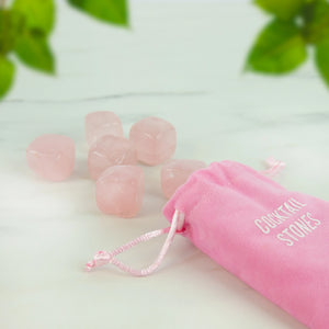 Set of six rose quartz cocktail stones laid out next to an open pink velvet pouch on a white marble background with green leaves.
