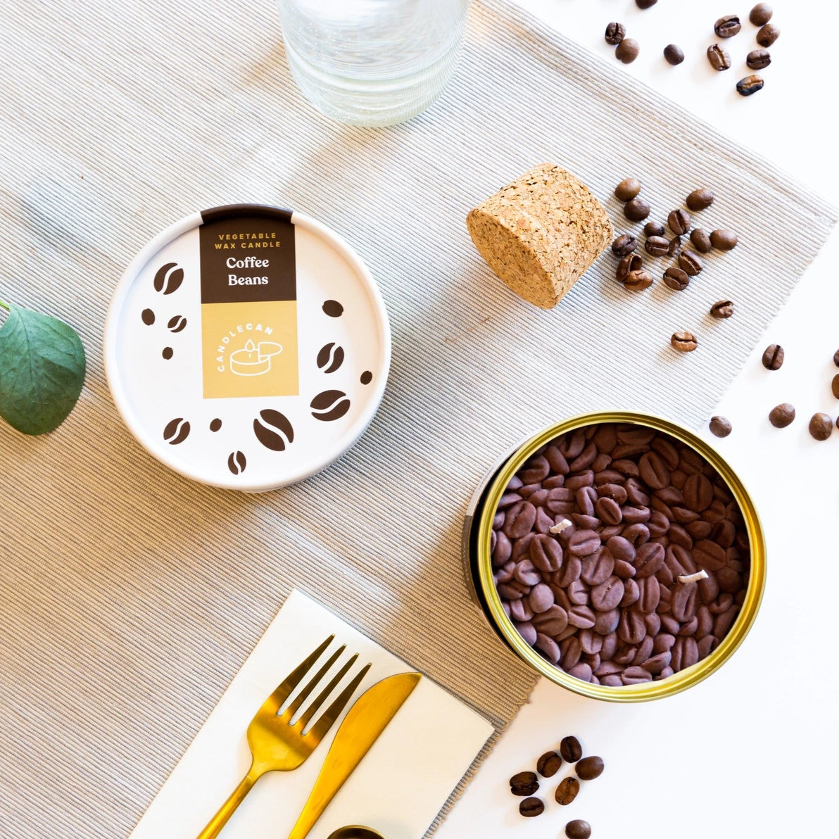 Coffee-scented candle styled on a table setting with eucalyptus, gold cutlery, scattered coffee beans, and the unopened Coffee CandleCan tin.