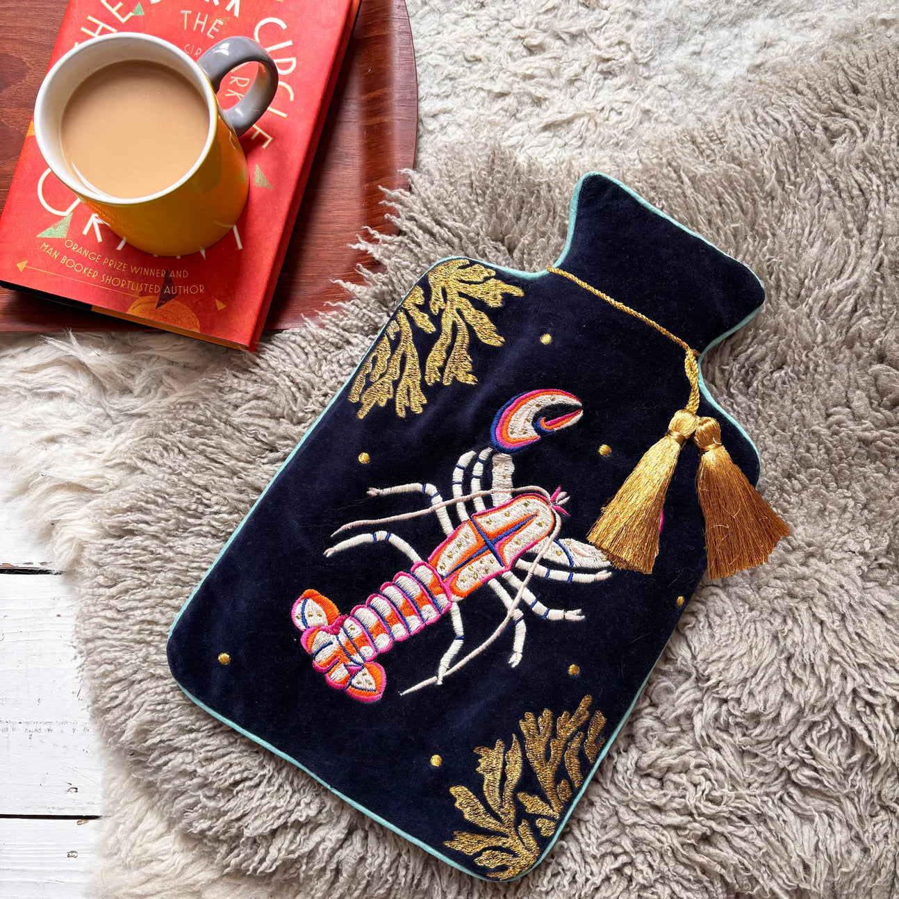 Coral Lobster Hot Water Bottle on faux fur rug with book and mug of tea – navy velvet embroidered with lobster and gold coral.