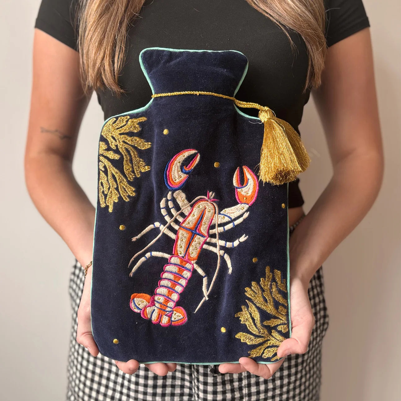 Woman holding Coral Lobster Hot Water Bottle – navy velvet with embroidered lobster design and gold tassel.