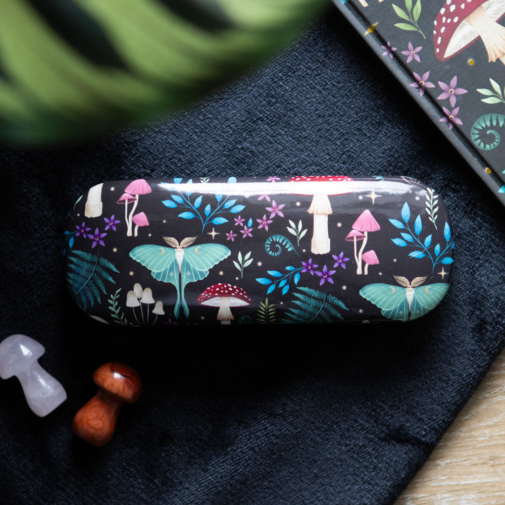 Dark Forest glasses case on black fabric with mystical mushrooms, moths and botanical print; leafy plant and matching notebook nearby.