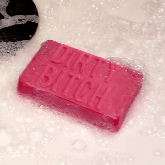 Pink “Dirty Bitch” soap lathered in bubbles floating in a bath or sink, surrounded by foam.