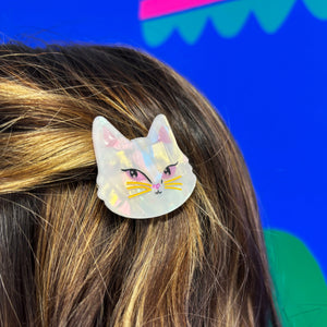 Close-up of a white cat-shaped hair clip attached to a person’s hair, featuring iridescent acrylic with golden whiskers and pink detailing.