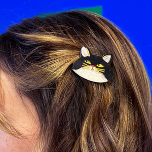 Close-up of a black cat-shaped hair clip in a person’s hair, featuring striking yellow eyes, golden whiskers, and white-tipped ears.