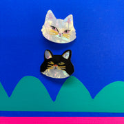 Flat lay of two cat-shaped hair clips—one white and one black—against a vibrant blue background with green and pink geometric accents.