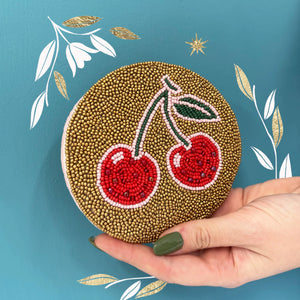 Round beaded coin purse with red cherry design and gold background held against teal wall with decorative leaf graphics.
