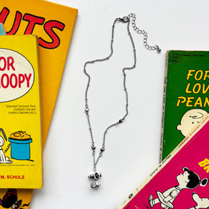 A silver Snoopy necklace displayed on a white background, surrounded by vintage Peanuts comic books.