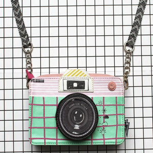 Front view of the Disaster Designs Pix Camera Mini Bag with a pink and mint green grid pattern, black lens design, and a playful lightning bolt charm.