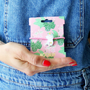 A person holding the Disaster Designs Snorkmaiden Enamel Hairband in its original pink floral packaging, against a denim background.