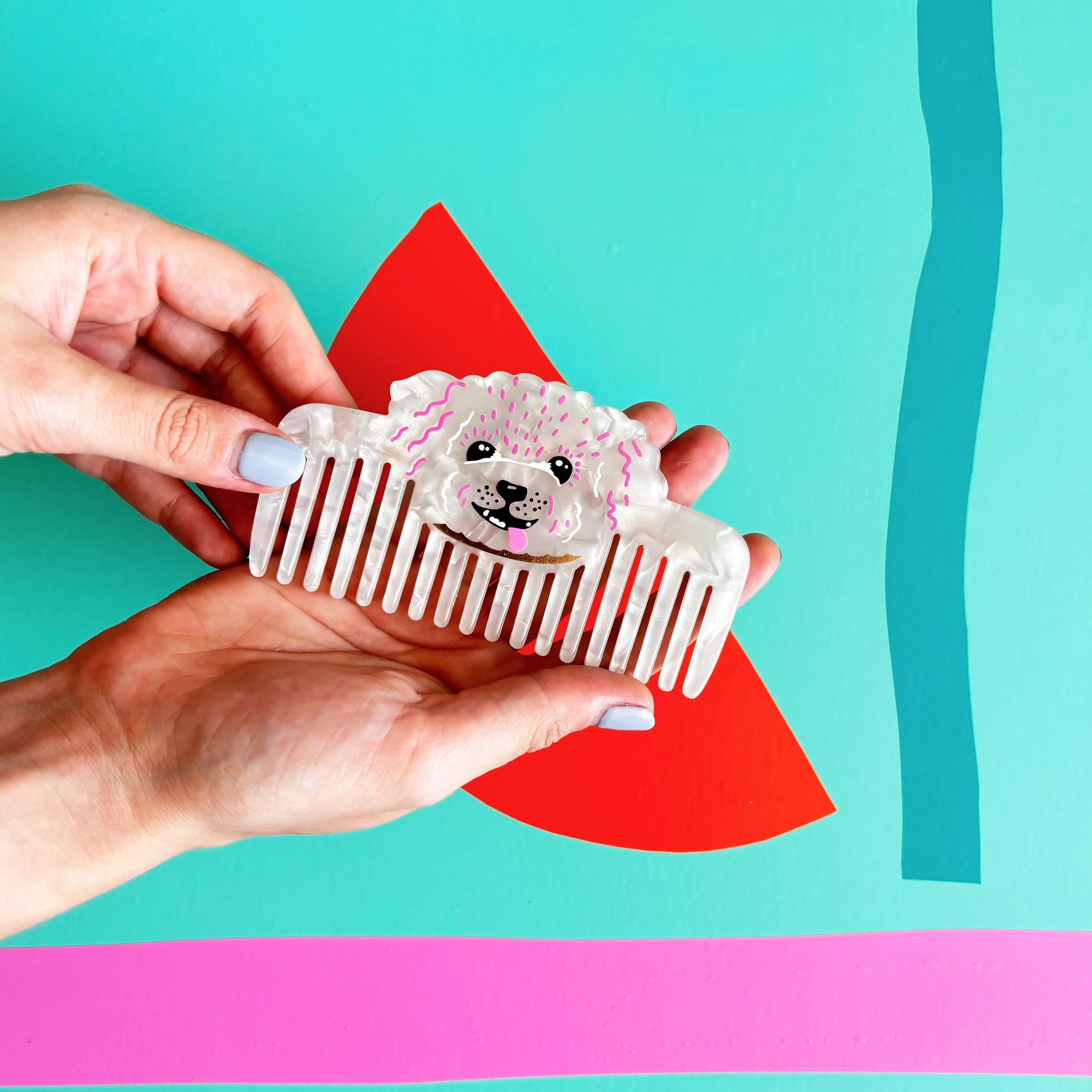Hands holding pearlescent dog-shaped comb against colourful background – Dog House Hair of the Dog Comb.