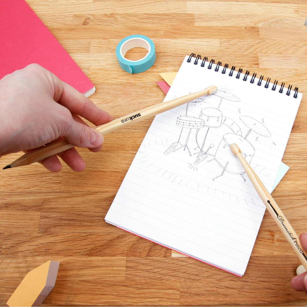 Person using wooden drumstick pencils to sketch a drum kit in a notebook on a wooden desk, showing playful and creative use