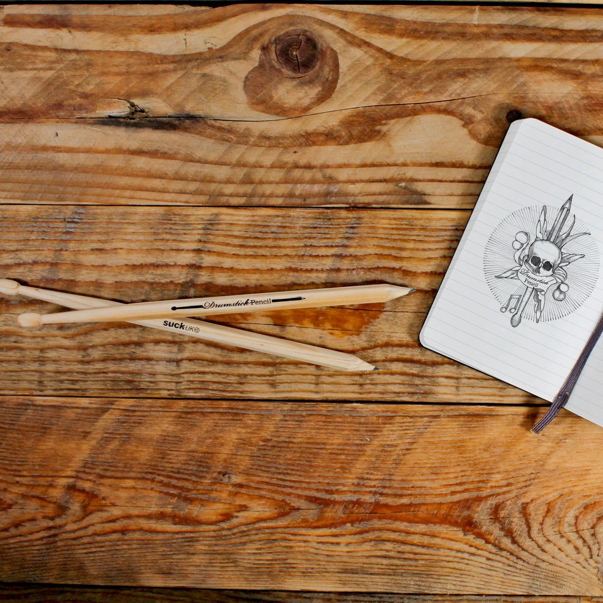 Drumstick pencils resting on a rustic wooden surface next to a notebook with a tattoo-style skull and feathers sketch