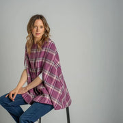 Woman wearing a purple plaid poncho sitting on a stool against a gray background. Earth Squared Winton Tweed Cape in bold pink check.