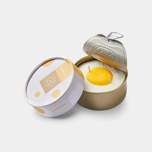 Vanilla Egg Candle in open tin with peeled-back lid, showing a realistic sunny-side-up egg design in creamy white wax, packaged in a round illustrated container.