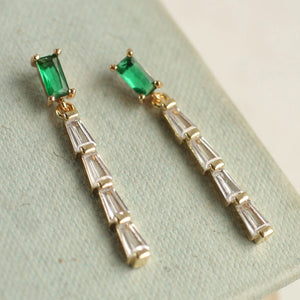 Another close-up of the emerald baguette crystal drop earrings placed on a textured background. The earrings highlight their intricate gold-tone setting and shimmering clear stones.