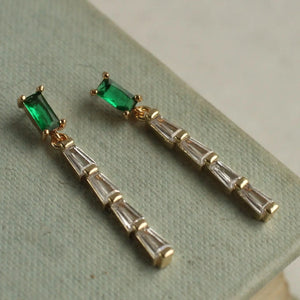 A pair of emerald baguette crystal drop earrings displayed on a textured light green surface. The earrings feature a delicate Art Deco-inspired design with emerald green and clear diamond-cut stones.