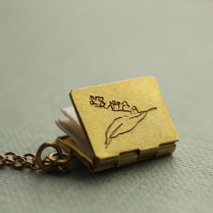 A delicate brass book locket with an engraved lily of the valley flower, partially open to reveal folded pages inside, lying on a soft green background.