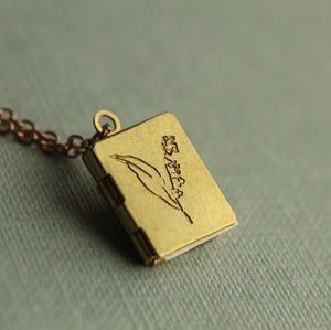 A brass book locket with an intricate lily of the valley engraving, hanging from a vintage-style chain, placed on a muted green fabric surface.