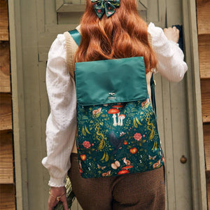 Lifestyle Image: A model wearing the Into the Woods Backpack, standing in front of a rustic wooden door, styled with a matching green bow in her hair.