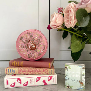 The pink dormouse jewellery box displayed on a stack of classic books with a floral arrangement in the background, creating a charming, vintage aesthetic.