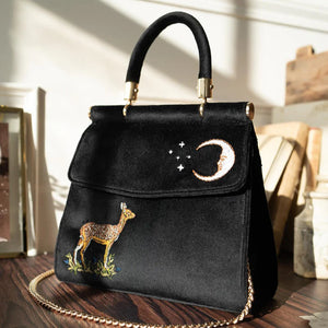 Lifestyle Shot: The tote bag placed on a wooden surface with soft lighting, highlighting the texture of the velvet and the detailed embroidery of the deer and celestial design.