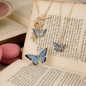  A styled shot of the Fable England Enamel Blue Butterfly Short Necklace, displayed alongside other butterfly-themed jewellery on an open vintage book. 