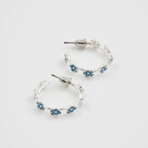 Angled overhead shot of silver hoop earrings with blue forget-me-not enamel flowers in a natural twig-inspired design.