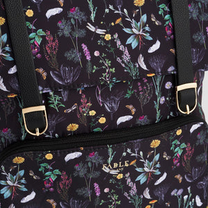 Close-up of gold Fable England logo and zip on front pocket of black floral backpack with detailed botanical pattern.