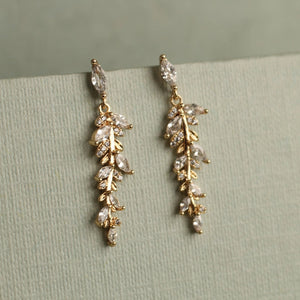 A pair of gold-plated feather fern drop earrings adorned with clear, diamond-like stones, displayed against a muted green textured background.