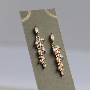 Elegant feather fern drop earrings with sparkling stones, presented on an olive green display card with gold detailing.