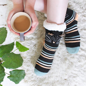 Cosy Feline Slipper Socks with cat face design and fleece lining, shown on feet beside a cup of tea on a fluffy rug.