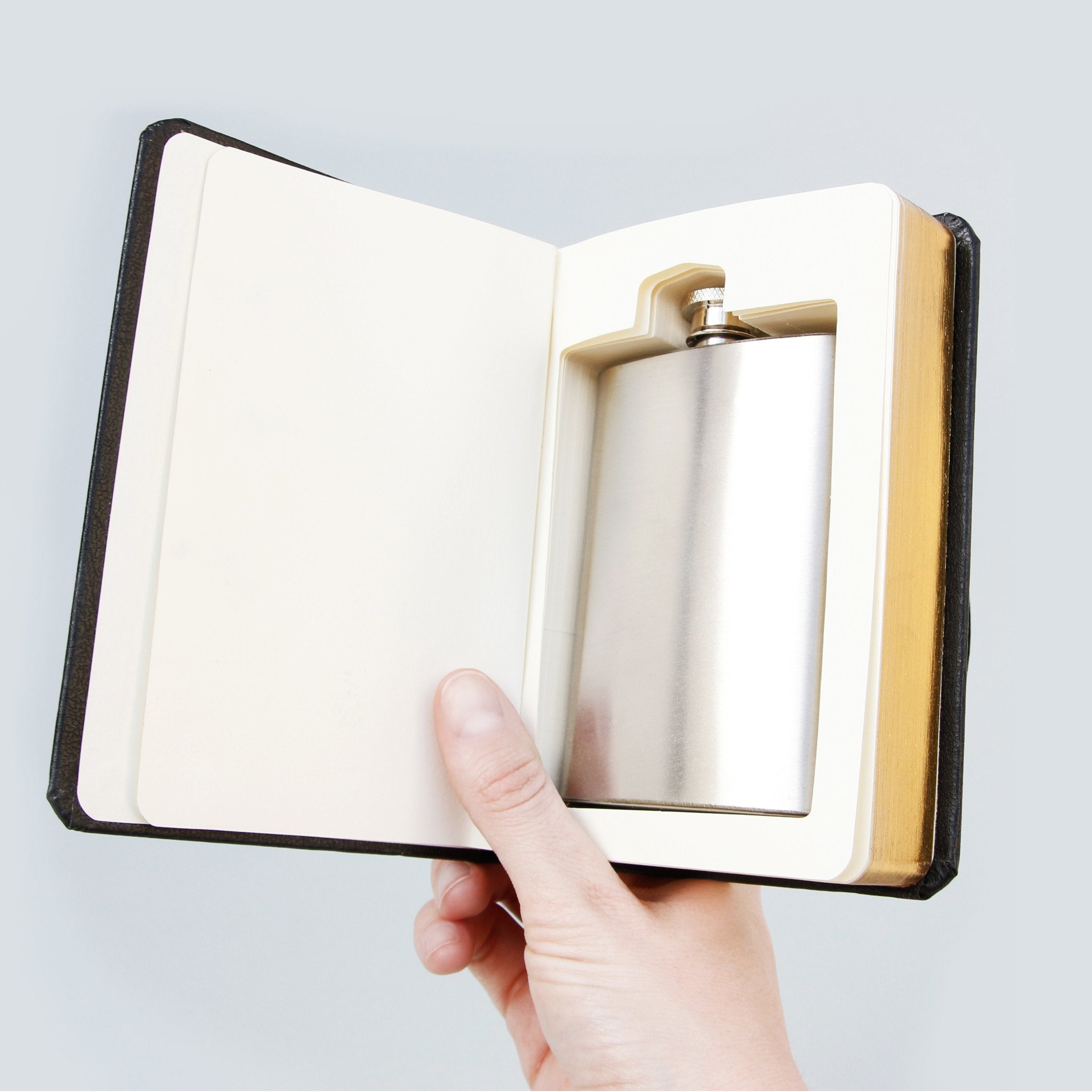 Hand holding open “Good Book” novelty flask gift, revealing the concealed stainless steel hip flask fitted snugly into the carved book interior.