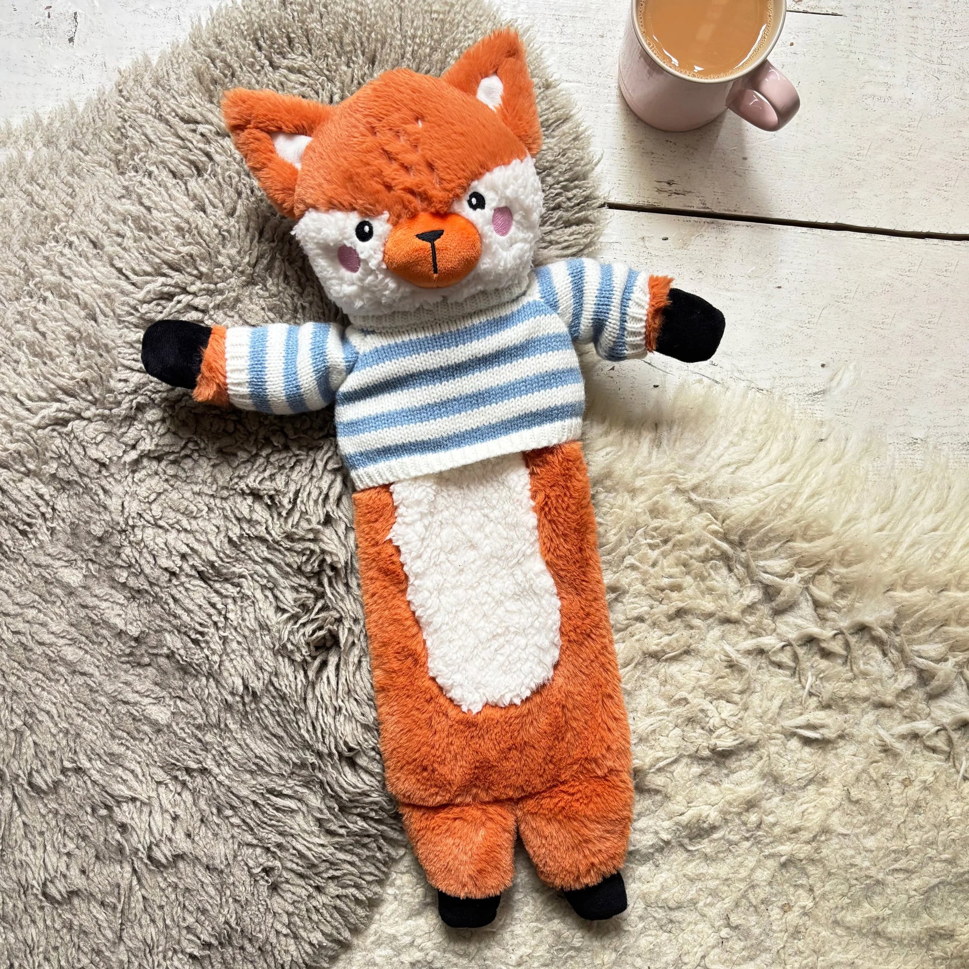 Fox Hot Water Bottle with striped jumper lying on sheepskin rug beside a cup of tea.