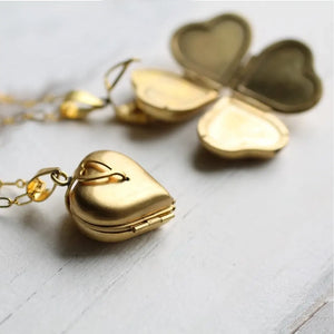 A gold heart-shaped locket with an ornate clasp, opened to reveal four spaces for photos, lying on a white wooden surface.