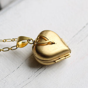 A close-up of the closed gold heart-shaped locket with a delicate gold chain, featuring a small heart-shaped detail on the clasp.