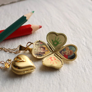 The Friends & Family Locket fully opened, displaying four heart-shaped photo slots with pictures and handprint designs inside, next to coloured pencils on a white wooden surface.