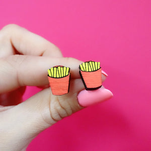 Hand holding a pair of fries stud earrings made from FSC-certified wood, painted in red and yellow with a fast food design against a bright pink background.