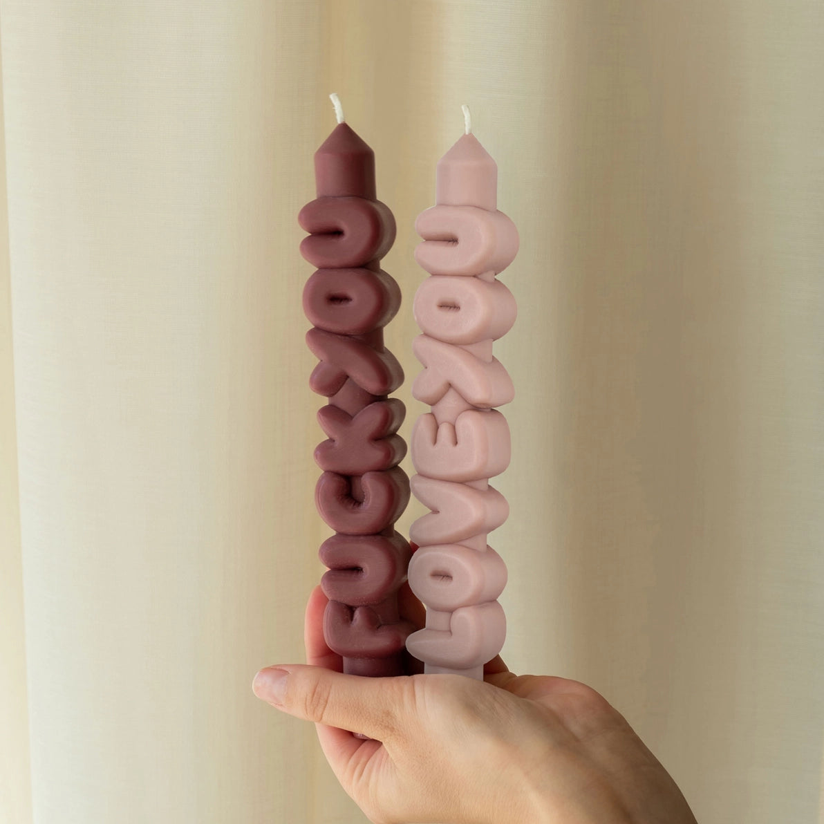 Mocha and cream statement candles reading ’Fck You’ and ‘Love You’, shown from the side and held in hand against a neutral curtain backdrop.
