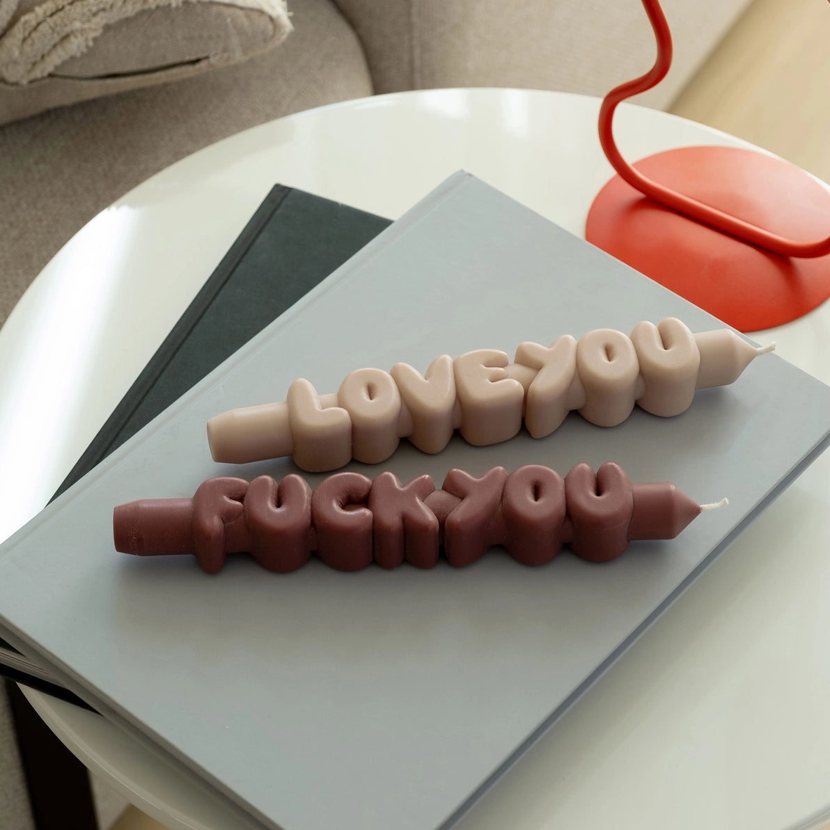 Playful ’Fck You’ and ‘Love You’ taper candles in mocha and cream colours displayed horizontally on a grey book, styled on a modern coffee table.
