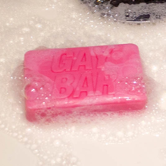 Pink “GAY BAR” soap lathered with bubbles in a sink – playful rose-scented novelty LGBTQ+ gift.