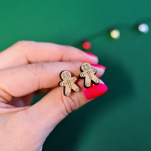 Close-up of a hand holding a pair of miniature wooden gingerbread man stud earrings with pastel icing detail, set against a festive green background.