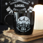 Black ceramic mug with ‘Girls Will Be Girls’ witch design on wooden board beside candle.