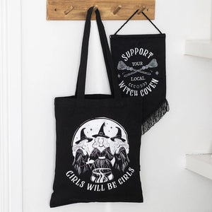 Black tote bag with ‘Girls Will Be Girls’ witch design hanging on wall.