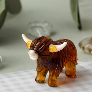 Glass Highland Cow Ornament on a table – A handcrafted glass Highland cow figurine with amber-toned glass, white horns, and intricate detailing, placed on a checkered cloth.