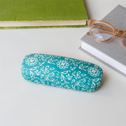 Front view of teal Radhika glasses case with white floral pattern, placed beside books and glasses..