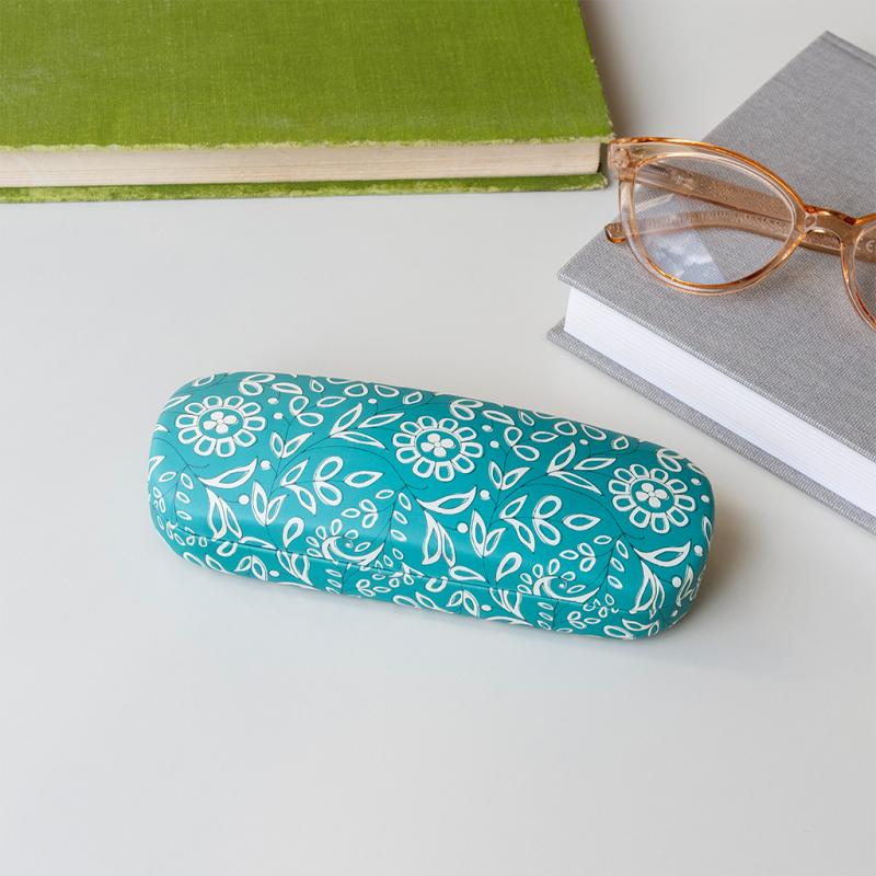 Front view of teal Radhika glasses case with white floral pattern, placed beside books and glasses..