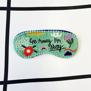Flat lay of Go Away I’m Busy velvet eye mask with embroidered text and colourful design.