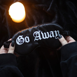 Black satin sleep mask with “GO AWAY” embroidered in white, shown on crumpled bedding with branded card – luxury eye mask with attitude.