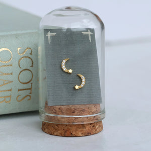 Moon and star stud earrings in a glass display cloche with cork base, styled next to a gold-embossed book.