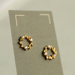 Gold-plated laurel wreath stud earrings displayed on a textured grey earring card, showing delicate leaf detailing and crystal accents.